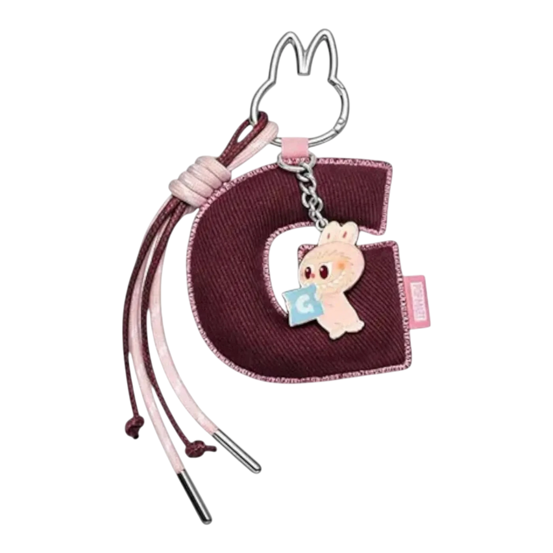 HypeSelection Labubu 'G' letter pendant keychain with pink bunny character holding a blue square. Burgundy fabric letter with pink and brown cords.